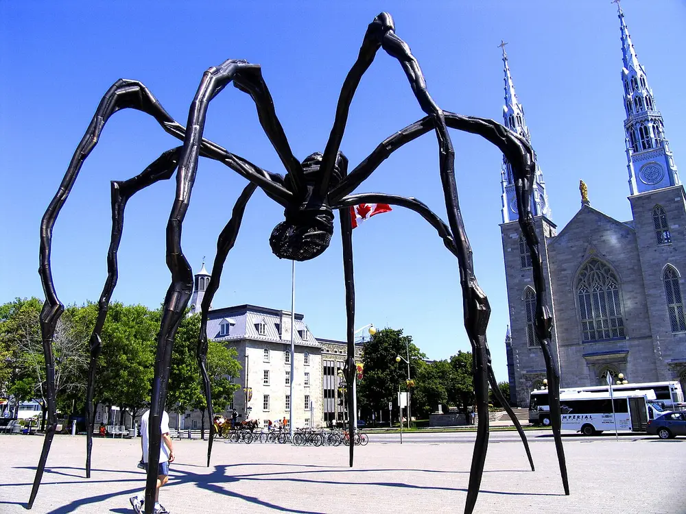 A giant bronze spider on thin, bending legs, taller than the buses and people below it, stands in a city square in front of a church with two tall spires.