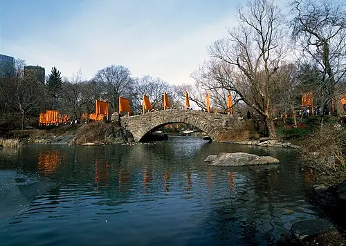 Rows of tall gates hung with saffron-orange fabric curve along a path past a stone bridge and a pond in Central Park in winter, among bare trees.