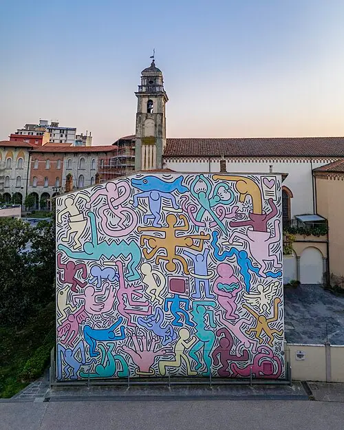 A large mural on the outside wall of a church, packed with brightly coloured cartoon-like figures with thick black outlines, linked together and dancing, under a bell tower.