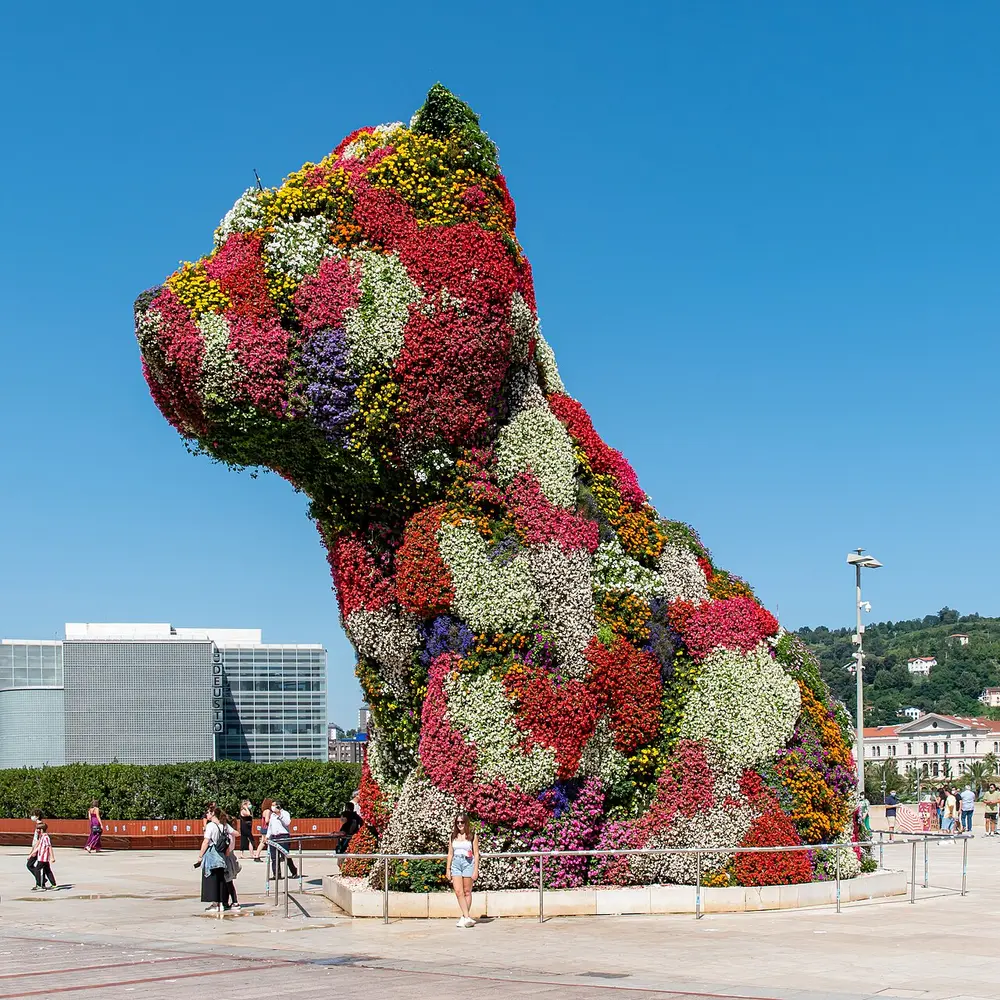 A giant sculpture of a sitting West Highland terrier puppy, covered all over in living flowering plants in red, pink and white, outside a museum under a blue sky.