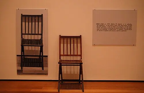 A gallery wall with a life-size black and white photo of a wooden folding chair on the left, the real chair standing in the middle, and a printed dictionary definition of the word chair on the right.