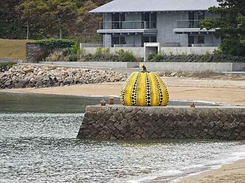 A large yellow pumpkin covered in rows of black dots sits on the end of a stone pier by the sea, with a beach and a modern building behind it.