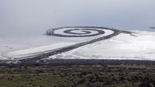 A long spiral path of dark rocks coils out into a pale, salt-white lake under a grey sky, seen from a hillside above the shore.