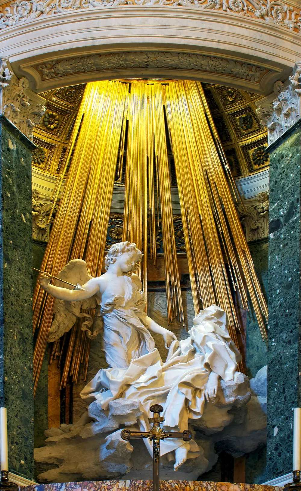 A white marble sculpture of a nun swooning on a cloud while a smiling angel holds a spear over her, in front of a burst of gilded rays, framed by green marble columns.