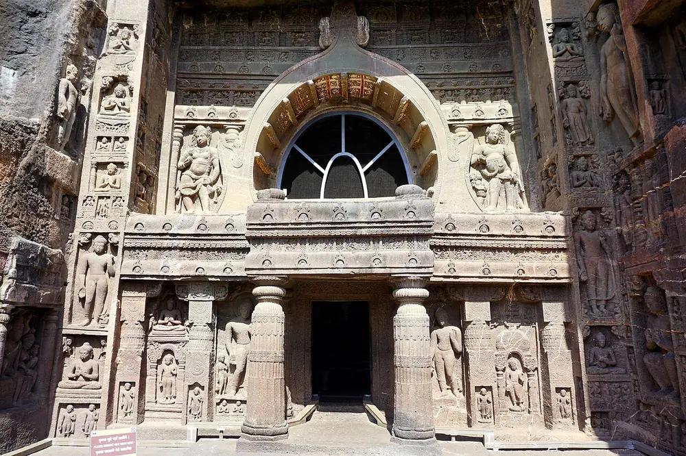 The carved front of Ajanta Cave 19: a large horseshoe-shaped window with a pointed top above a pillared porch, with figures of the Buddha carved in panels all over the cliff face around it.