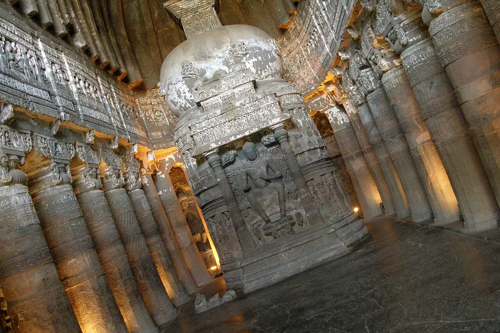 Inside a dimly lit rock-cut prayer hall: a tall stupa stands at the far end with a seated Buddha carved on its front, surrounded by rows of round, richly carved pillars and a ribbed ceiling.