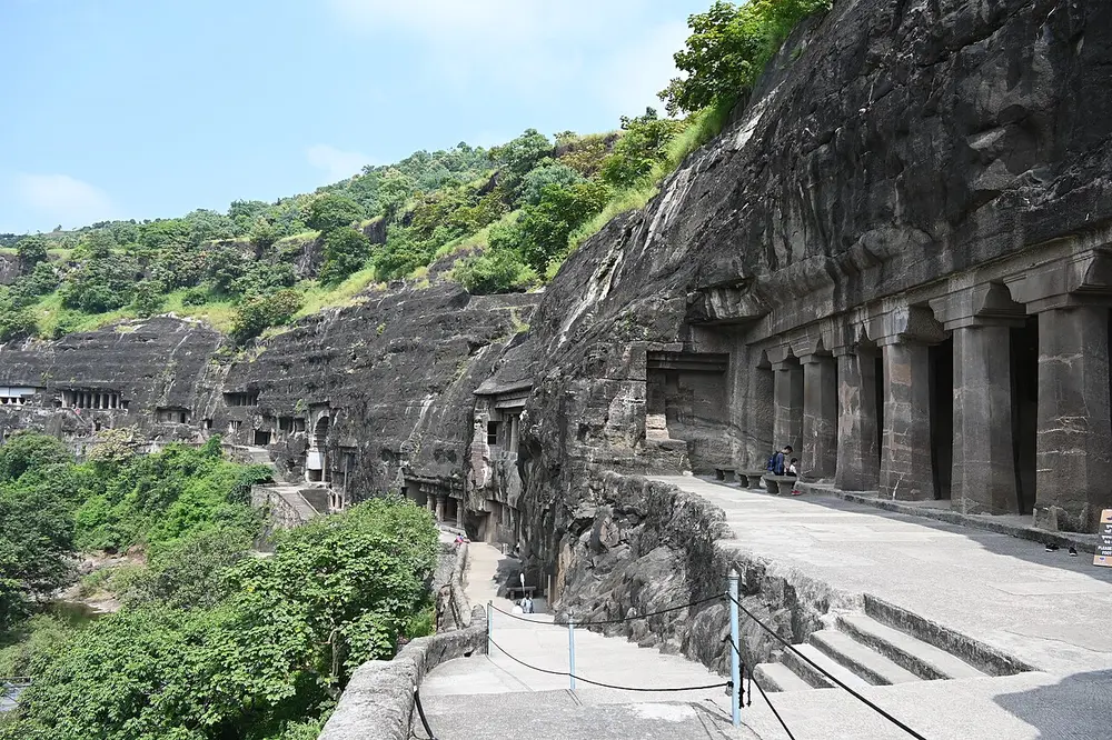 A walkway along a curving basalt cliff above a wooded river gorge; on the right, a row of thick square pillars marks the front of a large rock-cut monastery, and more cave doorways line the cliff in the distance.