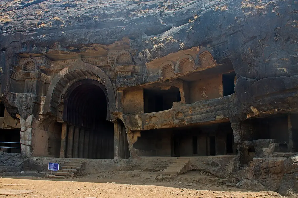 A dark basalt cliff with a tall open horseshoe-shaped arch leading into a vaulted hall, and rows of smaller rock-cut rooms and balconies beside it.