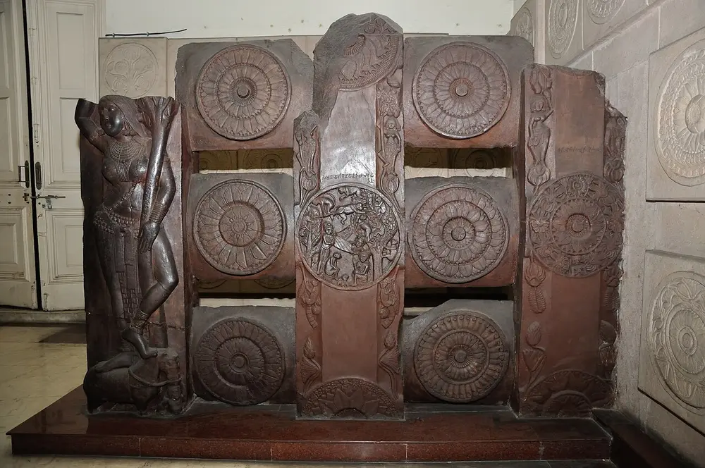 A section of an ancient red sandstone railing in a museum: upright posts and crossbars carved with big round lotus medallions, one medallion showing a story scene, and a tall carved woman holding a tree branch on the corner post.