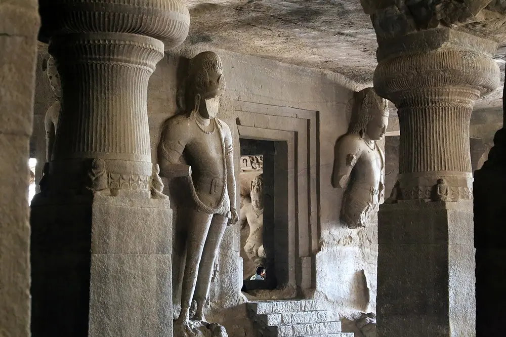 Inside a dark rock-cut hall: thick pillars with cushion-shaped tops frame a small square shrine, its doorway flanked by tall carved doorkeepers, with steps leading up to it.