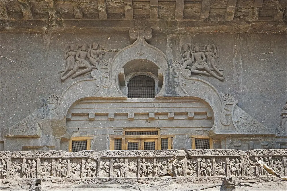 The carved upper front of a rock-cut hall at Ellora: a horseshoe-shaped window framed by a carved arch, flying couples carved on either side, and a balcony below with a frieze of small dancing figures.