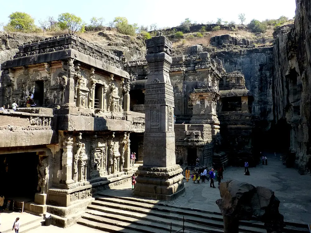 Looking down into a huge courtyard cut into a hillside: a two-storey carved temple stands on the left, a tall square stone pillar rises in the middle, and the main temple and the cut cliff walls rise behind.