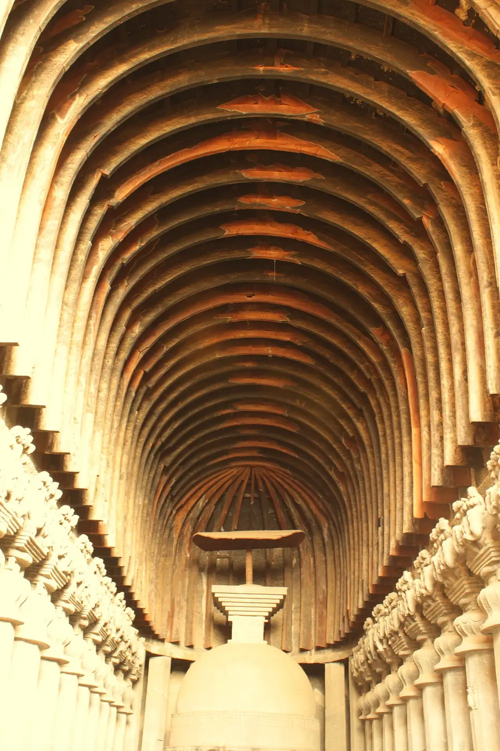 Looking down a long rock-cut hall towards a stupa: the high curved ceiling is lined with real wooden ribs, rows of pillars run along both sides, and a wooden umbrella stands on top of the stupa.