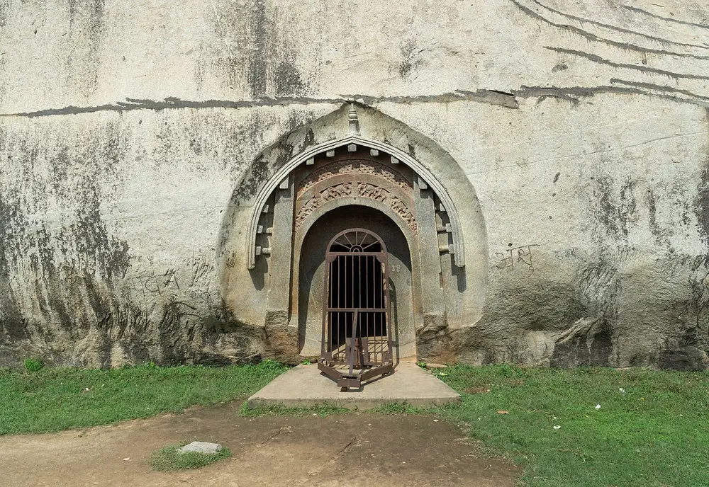 A doorway carved into a smooth grey granite cliff, shaped like the front of a hut with a pointed, curved roof, with a band of carved elephants over the door and an iron gate.