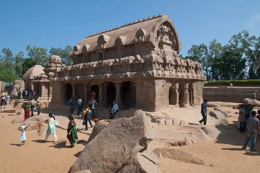 A long granite temple with a curved, barrel-shaped roof and pillared front, carved from rock and standing on sand, with smaller rock-cut temples behind it on the left and visitors walking around.