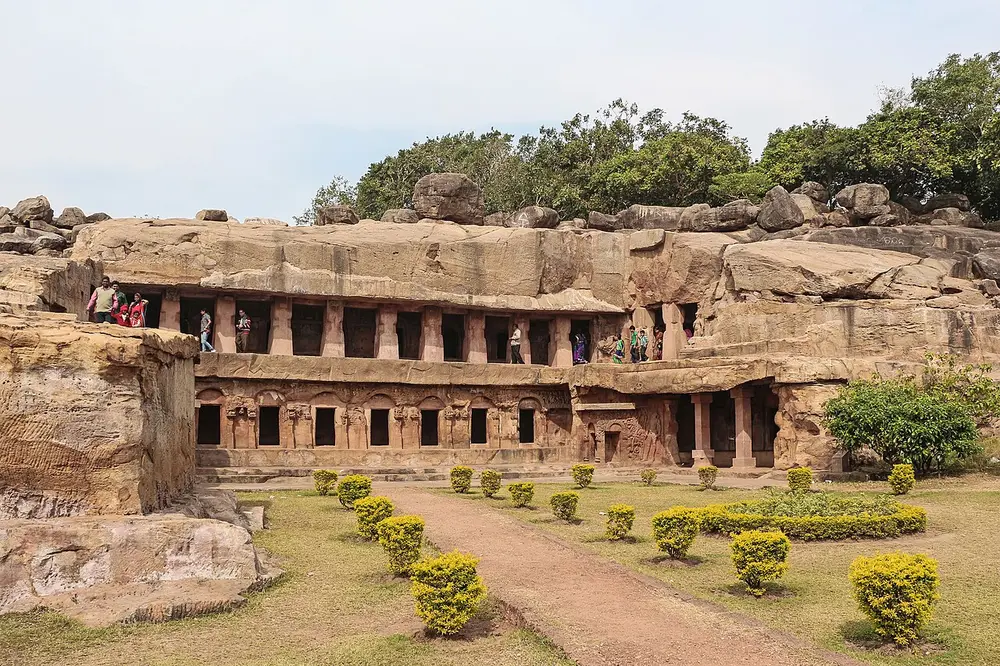 A two-storey row of rock-cut rooms in a low sandstone hill, with a pillared verandah on each level, carved friezes between the doorways, and a garden path in front.