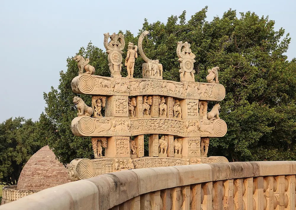 The top of a stone gateway at Sanchi: three curved stone beams with rolled ends, stacked one above the other on square posts, crowded with carved elephants, horses, riders and lions, with a wheel and figures standing on top.