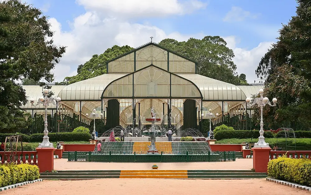 A Victorian glass house with arched roofs behind a fountain, in a garden with trees and lawns.