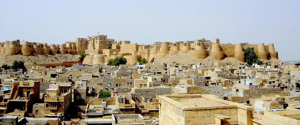 A long yellow sandstone fort with rounded towers on a hill, above a town of yellow stone houses.