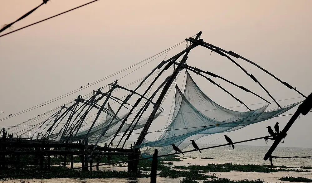 Large Chinese fishing nets on wooden frames, silhouetted against an evening sky by the sea.
