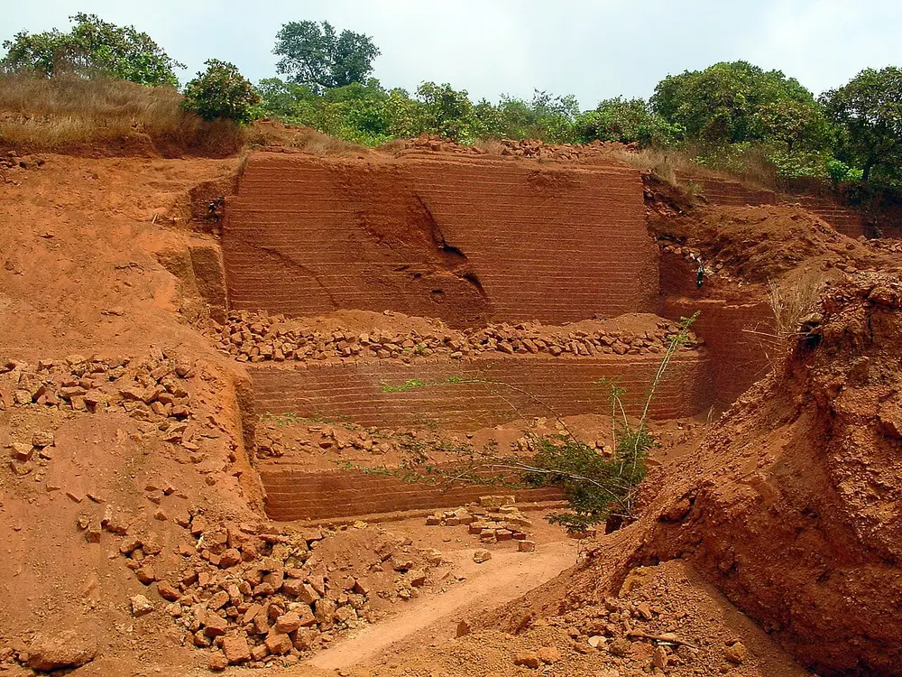A quarry with walls of bright red-orange earth cut into neat steps, with loose red blocks piled on each level.