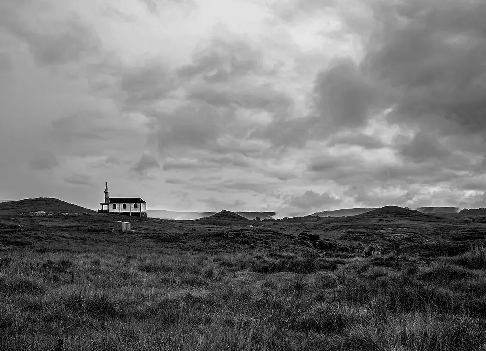 A black and white photo of a small white church on a grassy hill under heavy, rolling storm clouds.