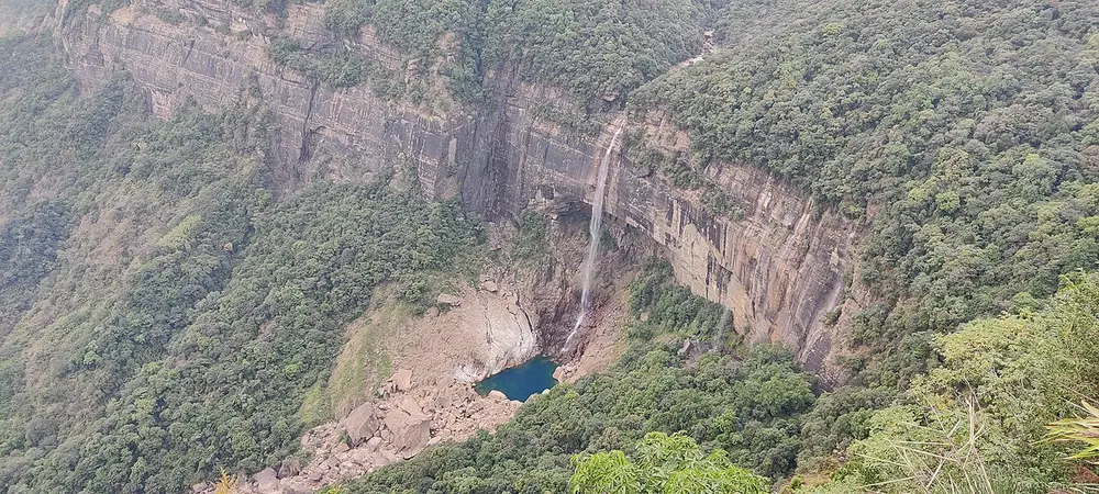 A tall, thin waterfall dropping down a sheer cliff into a small blue pool, surrounded by forested slopes.