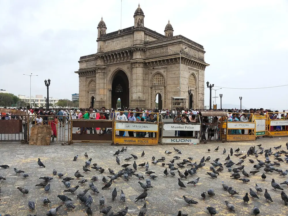 The Gateway of India, a tall yellow-brown stone arch with four turrets on top, seen behind a crowd and barriers, with pigeons in the foreground.