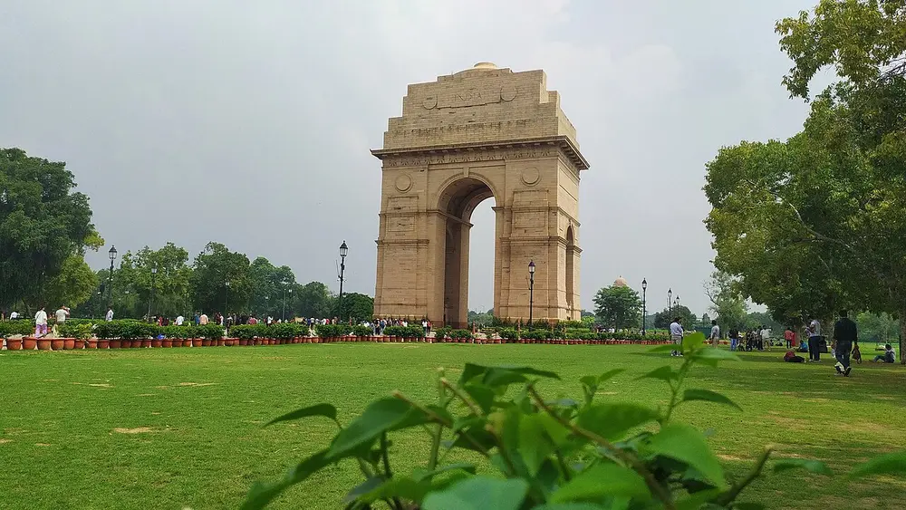 India Gate, a tall sandstone arch standing on a wide green lawn under a cloudy sky.