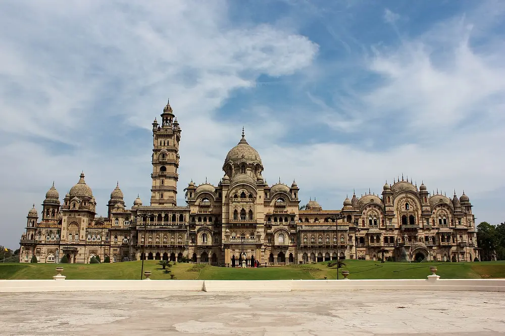 The long Laxmi Vilas Palace in Vadodara, a sandstone building with domes, turrets and a tall clock tower in the middle, behind a wide lawn under a cloudy sky.