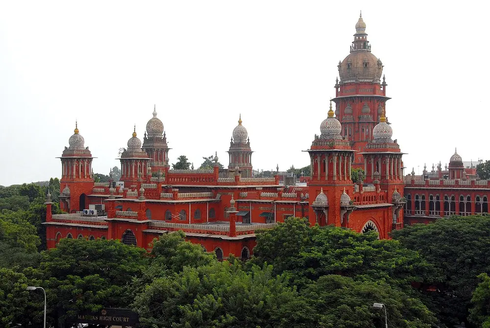The red brick Madras High Court with many domes and slim towers of different heights, the tallest topped by a large dome, above a mass of green trees.