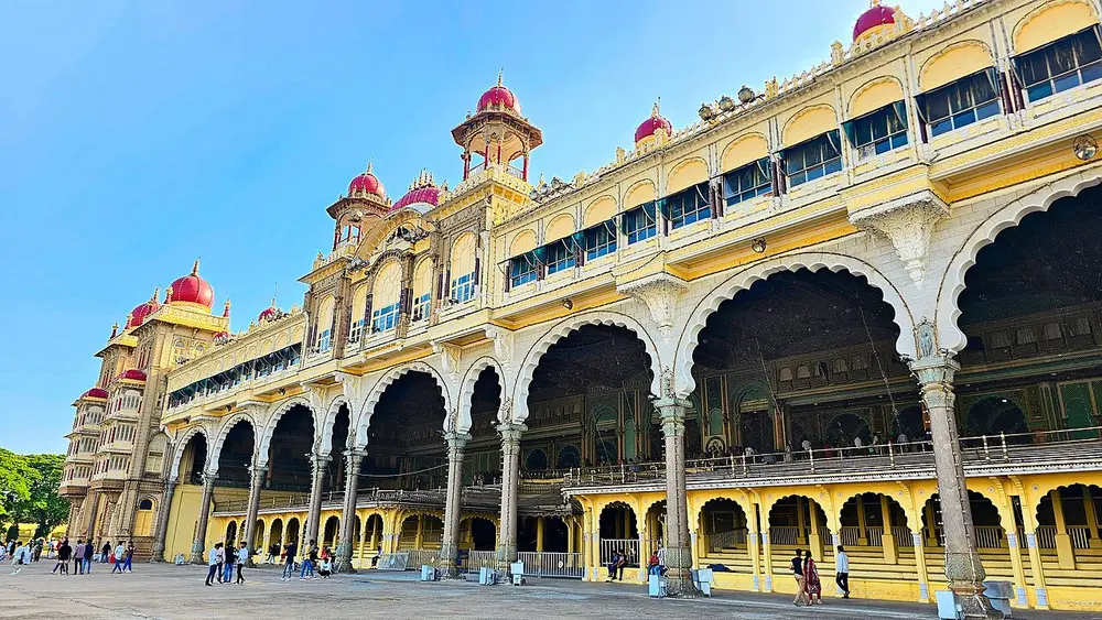 The front of Mysore Palace, a long cream and gold building with a row of big cusped arches on the ground floor, carved balconies above and red onion domes on its towers.