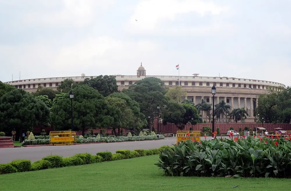 The round old Parliament House in New Delhi, a low circular building ringed by a long colonnade of pale sandstone columns, behind lawns and flower beds.