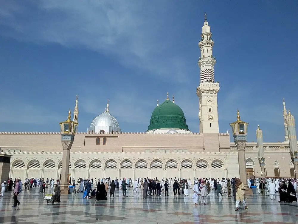 A wide white mosque courtyard with crowds of people, a tall pale minaret and a bright green dome under a blue sky.