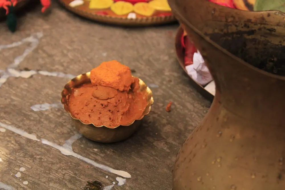 A small brass bowl heaped with orange-yellow turmeric paste, standing on a stone floor beside a large brass pot.