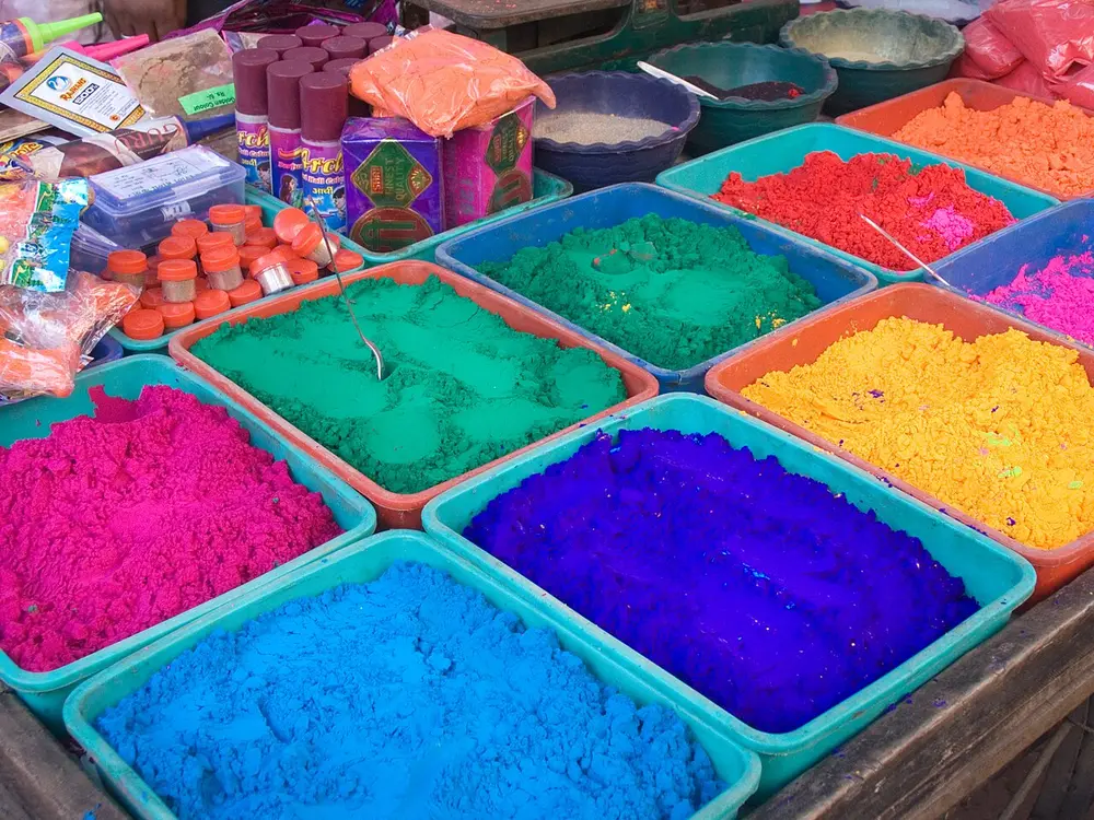 Square trays heaped with bright powder colours, pink, green, blue, purple, yellow and orange, on a market stall, with packets and squeeze bottles behind them.