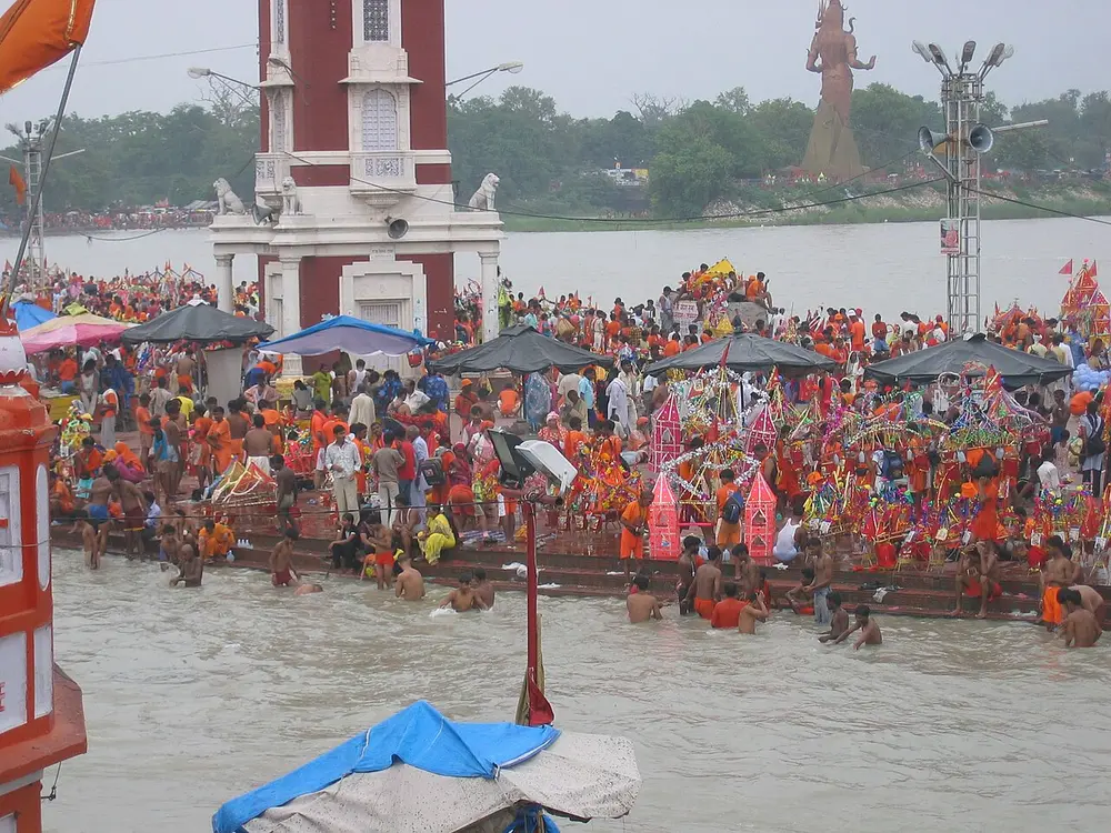 A huge crowd of pilgrims, most of them dressed in saffron and orange, fills the riverside steps and shallow water at Har ki Pauri in Haridwar, with a red clock tower behind them.