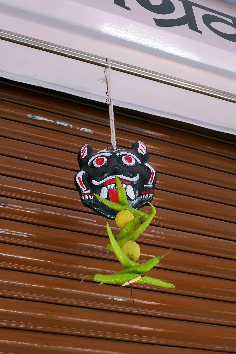 A small black demon-face mask with white eyes, fangs and a red tongue hangs above a string of green chillies and a lemon, in front of a brown shop shutter.