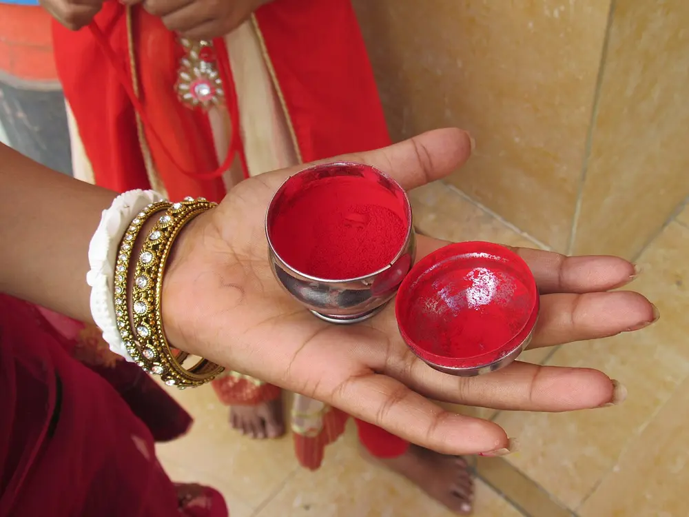 An open hand, with gold and white bangles on the wrist, holding a small round silver box and its lid, both filled with bright red powder.
