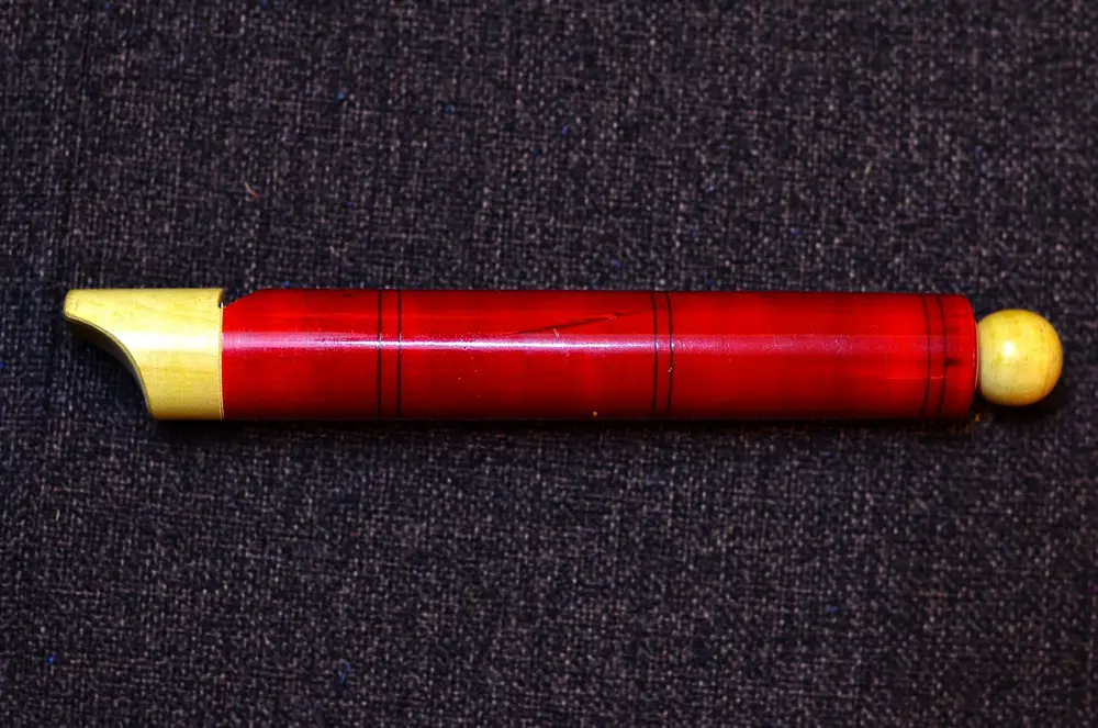 A red toy flute made of turned wood, glossy with lacquer, with a pale mouthpiece and a round knob at the end.
