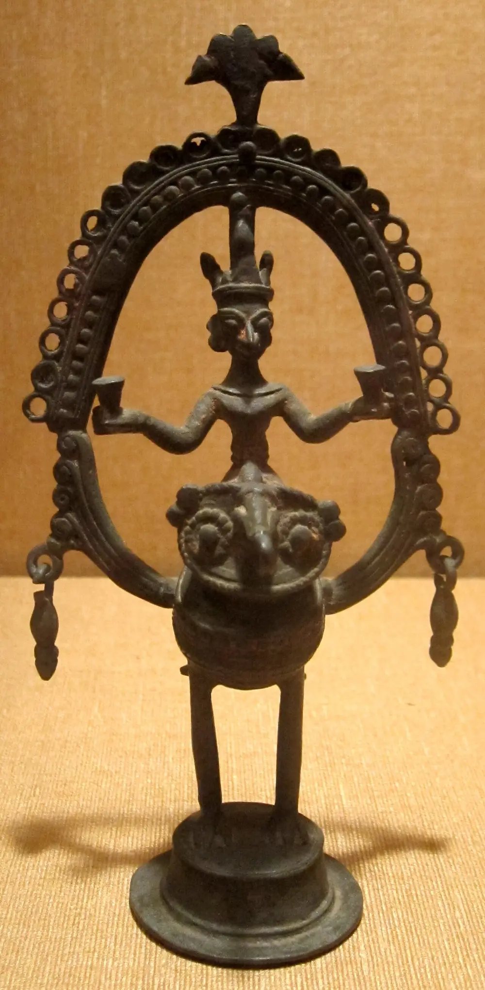 A dark brass figure of a god standing on an owl, framed by a tall arch of fine twisted metal threads with a leaf-shaped top.