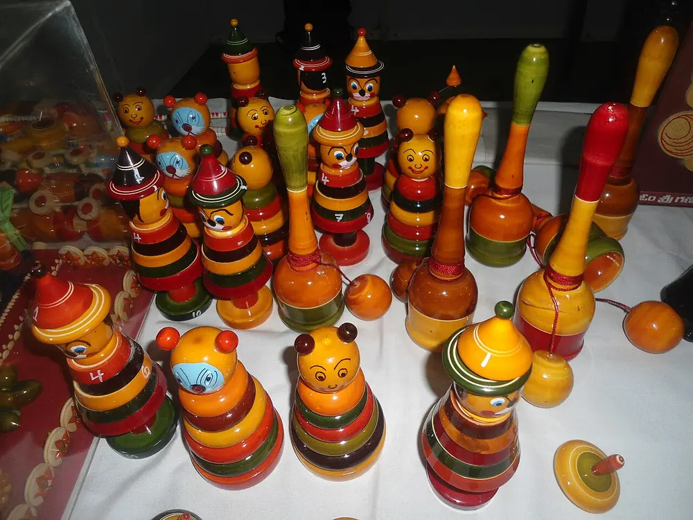 A table of glossy turned wooden toys in red, yellow, green and orange, including stacking dolls, tops and rattles.