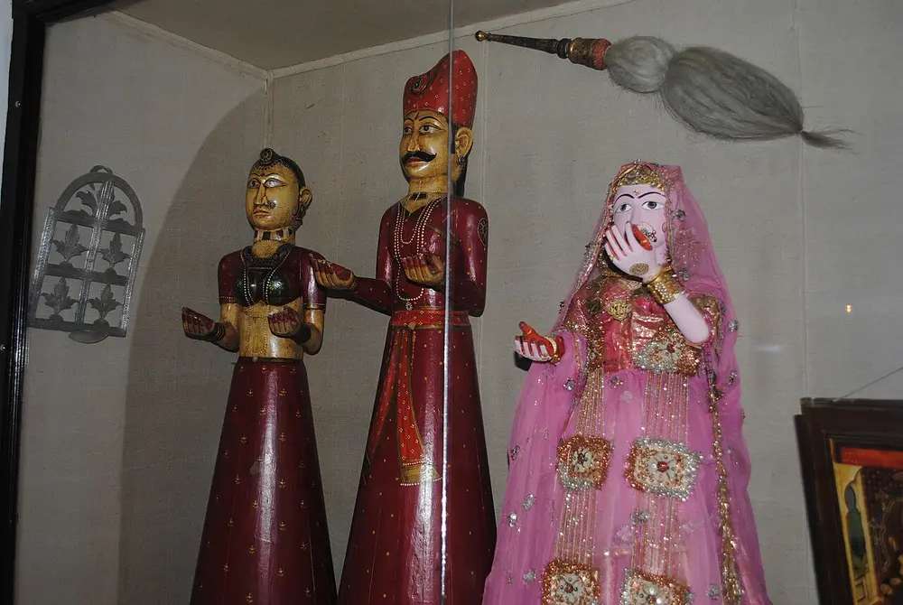 Three wooden string puppets in a museum: a woman in a red skirt, a man with a turban and moustache holding up a fly whisk, and a woman in a pink veil.