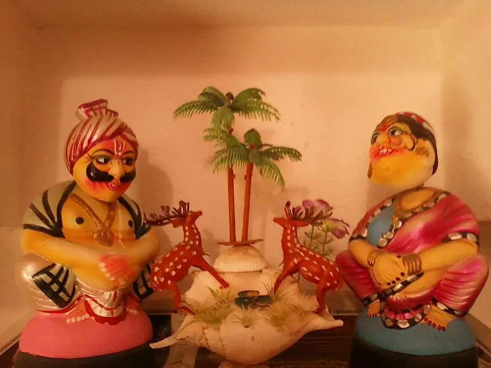 Two painted wooden figures, a man in a turban and a woman in a sari, sitting on either side of small palm trees and spotted deer.