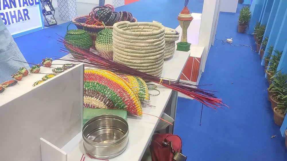A craft fair stall with coiled grass baskets and boxes in natural gold, and bundles of grass dyed bright red and green.