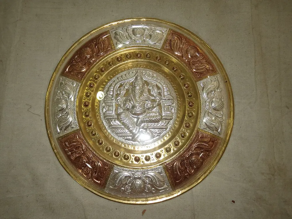 A round brass plate with a raised silver figure of a god at the centre, surrounded by rings and panels of embossed silver and copper.
