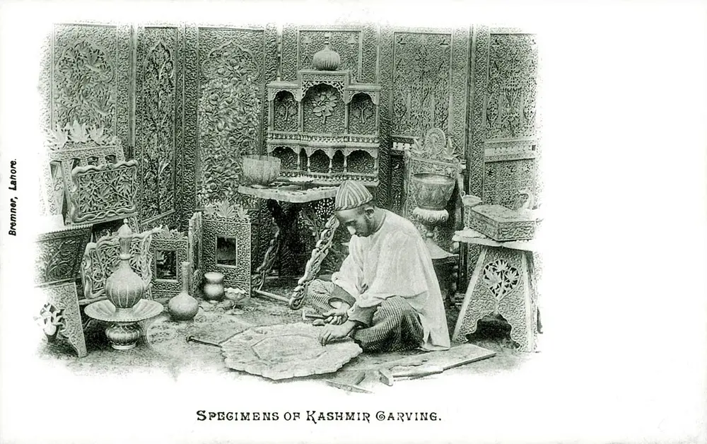 An old black and white postcard of a seated carver at work, surrounded by heavily carved screens, cabinets, tables and boxes.