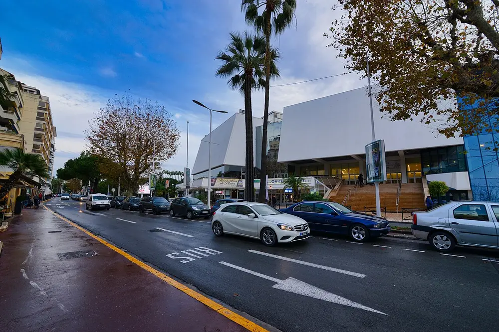 A large white modern building with slanted walls at the end of a palm-lined street, with parked cars and a wet road in front.