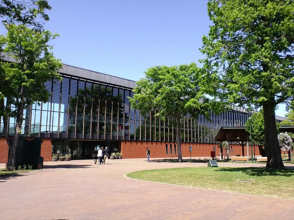 A long, low station building with a glass front in a grid of thin frames above a brick base, seen across a paved square with trees.