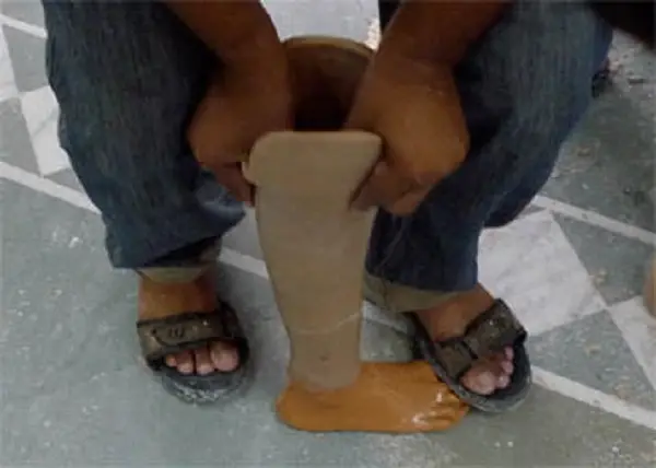 A seated person holds a beige artificial lower leg and foot, made of rubber, upright between their sandalled feet.